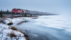 A passenger train in the distance traveling through tundra toward the icy shoreline of Hudson Bay near Churchill, Manitoba, with boreal forest fading into the Arctic landscape.