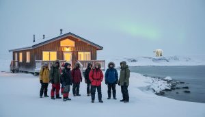Travelers stand outside a warmly lit tundra lodge in snowy Churchill, Manitoba, with a polar bear visible in the distant Arctic landscape.